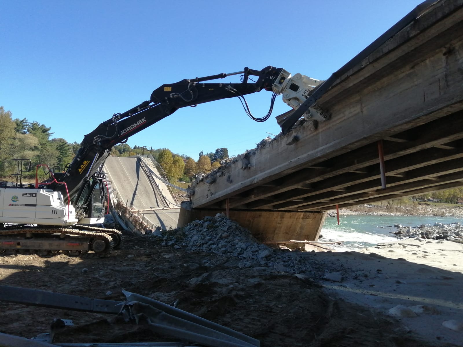 Un escavatore con un accessorio idraulico demolisce un'ampia sezione di un ponte in cemento armato vicino a un fiume.