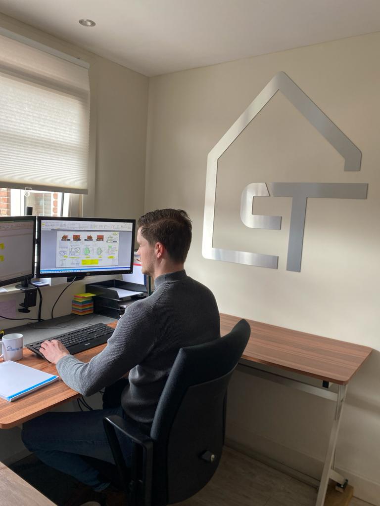 Iemand zit aan een bureau te werken op een computer in een kantoor met een groot, metalen, huisvormig logo aan de muur.