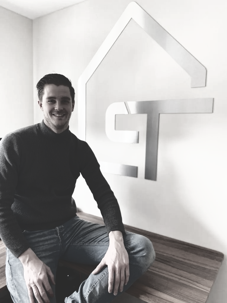 Een persoon zit glimlachend aan een bureau voor een muur met een groot, metalen, huisvormig logo met de letters ST.