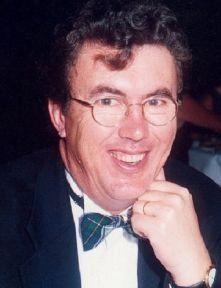 Man wearing glasses and a bow tie smiles, resting chin on hand.