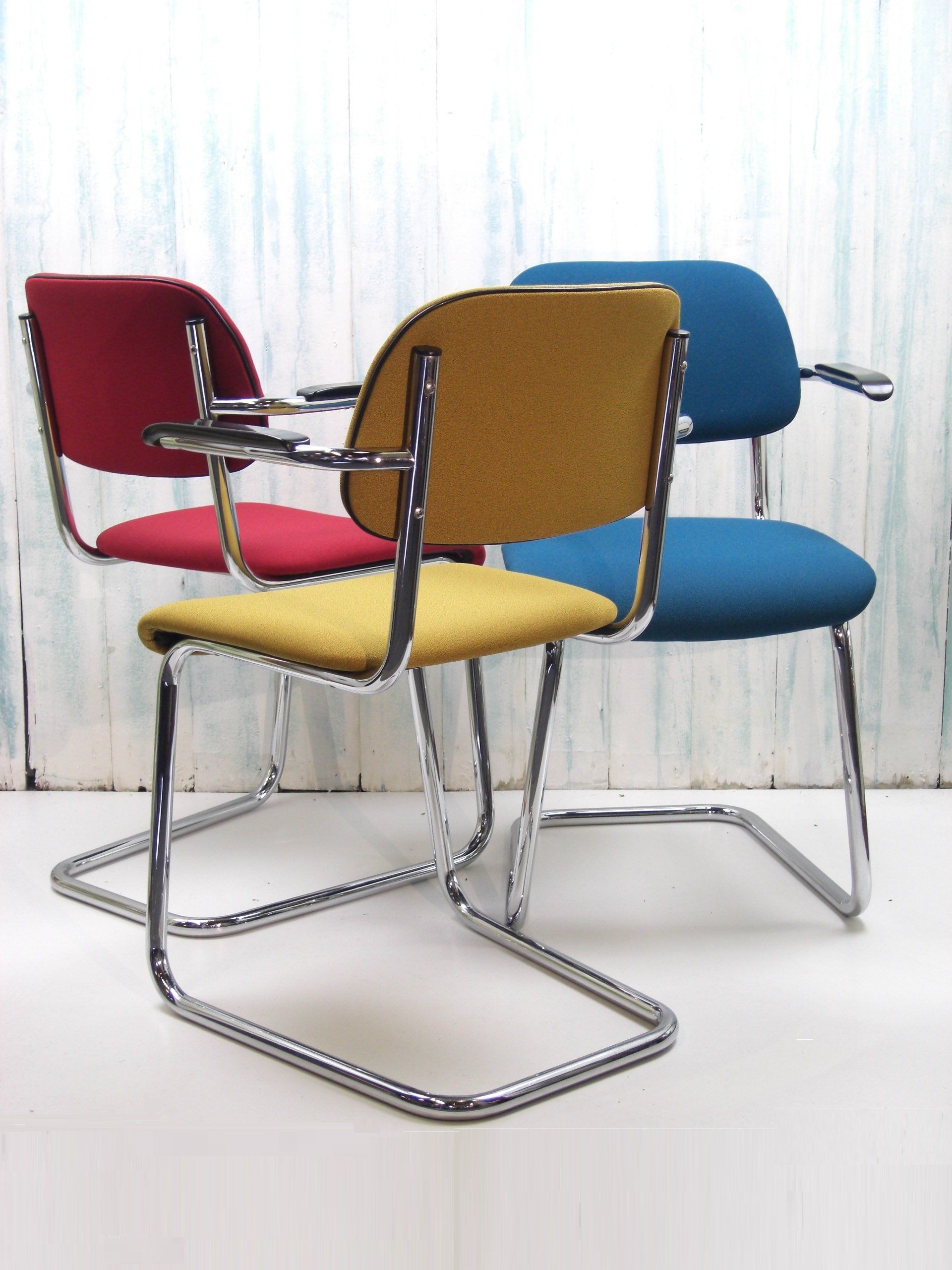 Drie kleurrijke cantileverstoelen: rood, geel en blauw, met chromen frames, geplaatst tegen een lichte, gestructureerde achtergrond.