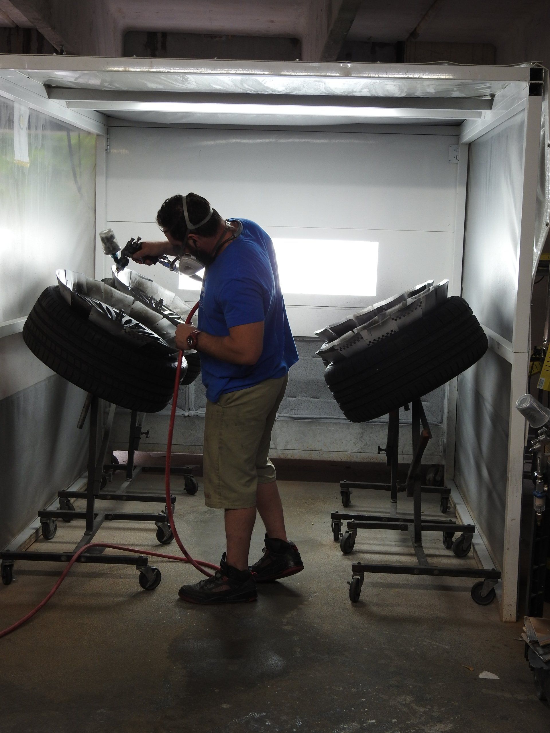 Man spraying tires in a paint booth. He wears a mask and khaki shorts. Two tires are on stands.
