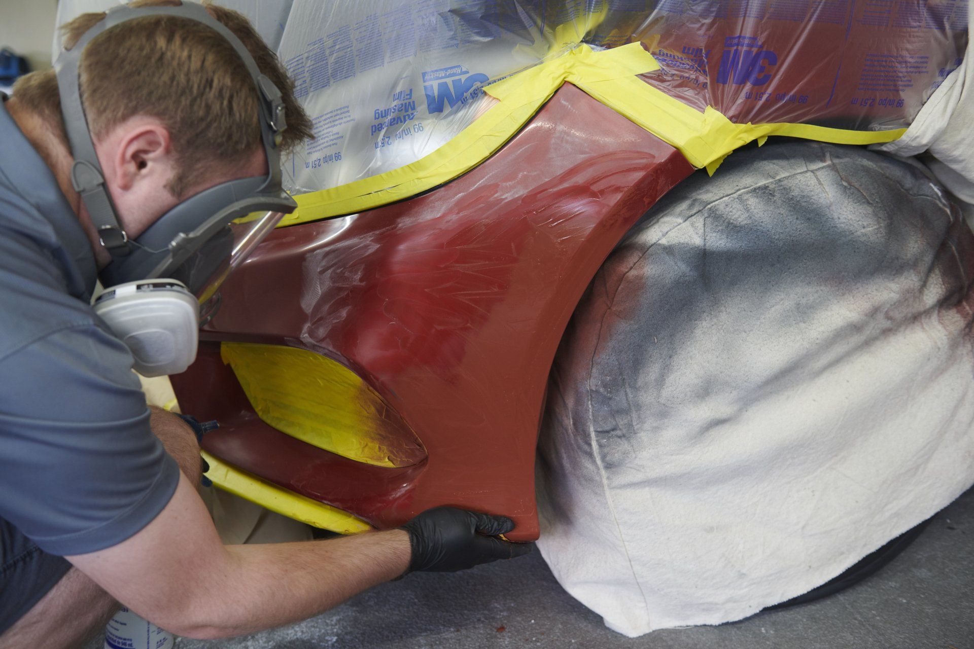 Person wearing a respirator working on a car bumper in a workshop, masked and taped, preparing for paint.