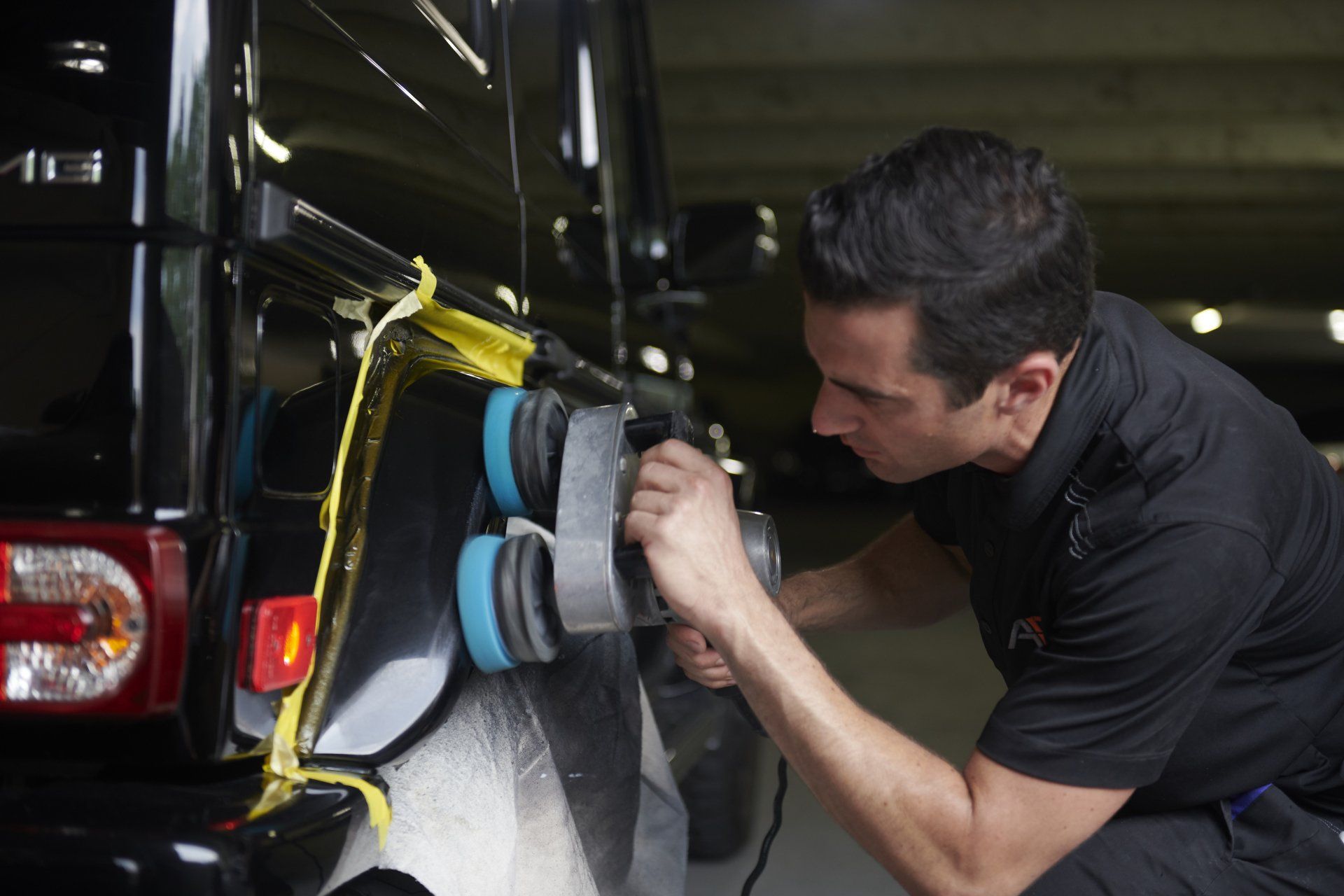 Person using a power buffer on a black car bumper, with protective tape around the edges.