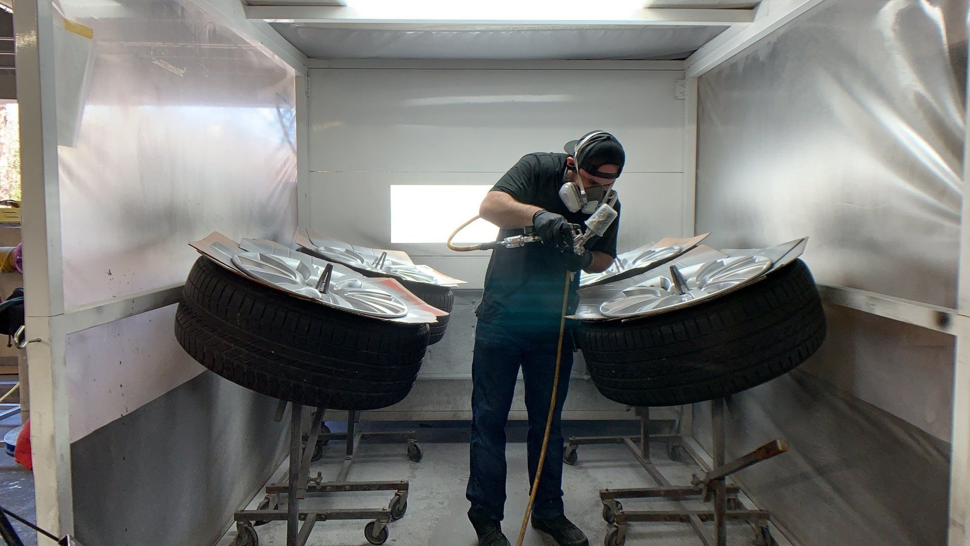 Person refinishing wheels inside a spray booth, two metal panels resting on tires, safety gear visible.