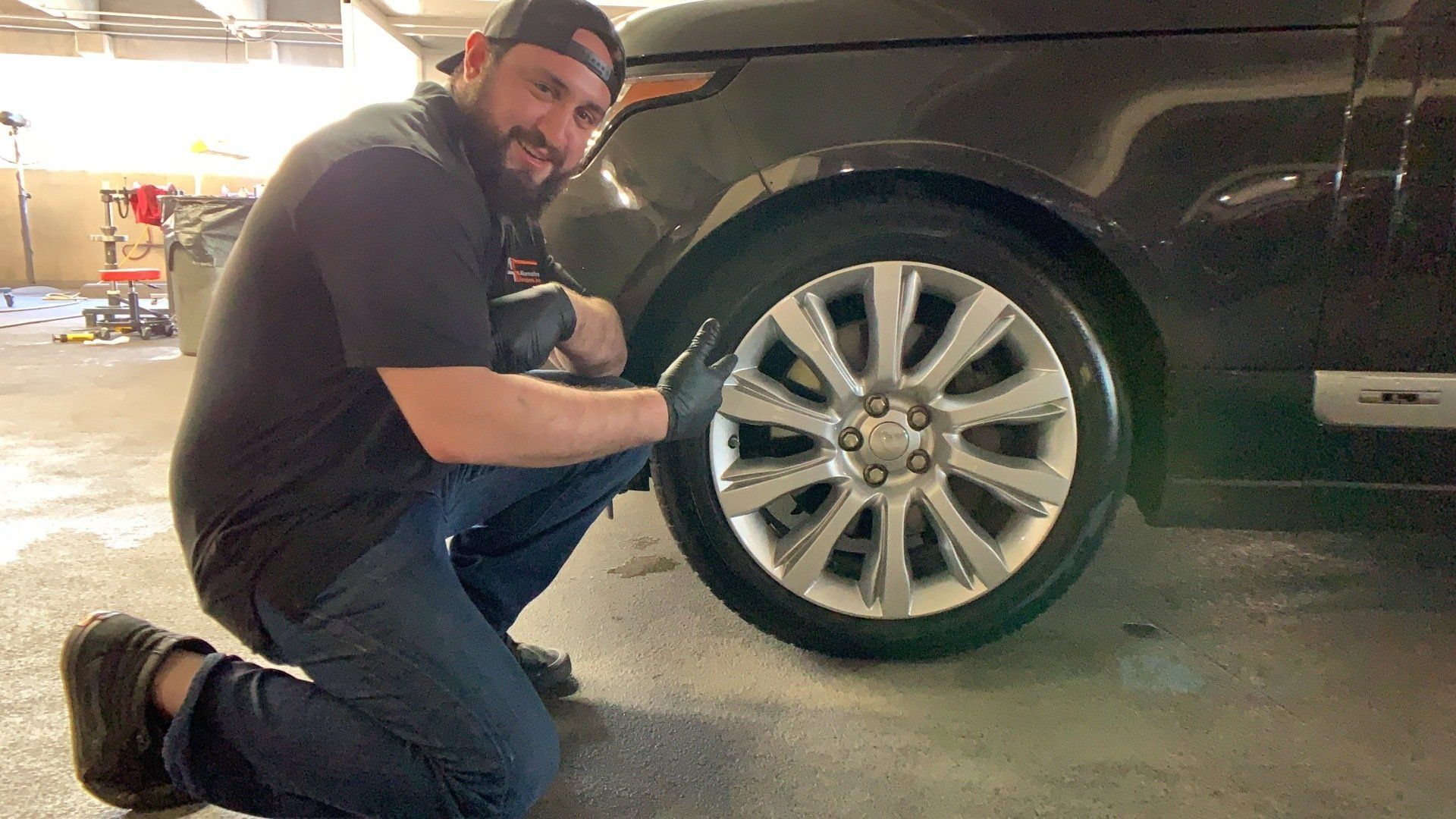 Man kneeling, pointing at car tire. He's wearing a hat, dark shirt, and gloves. Car is black, setting is a garage.