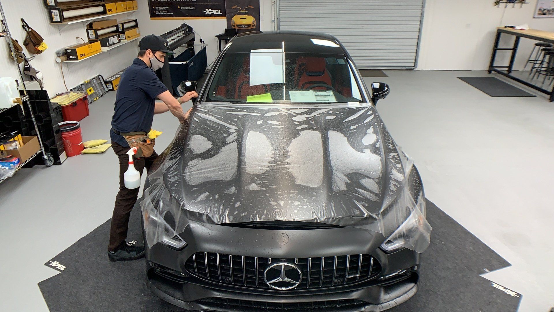 Person applying protective film to the hood of a dark grey Mercedes-AMG in a shop.