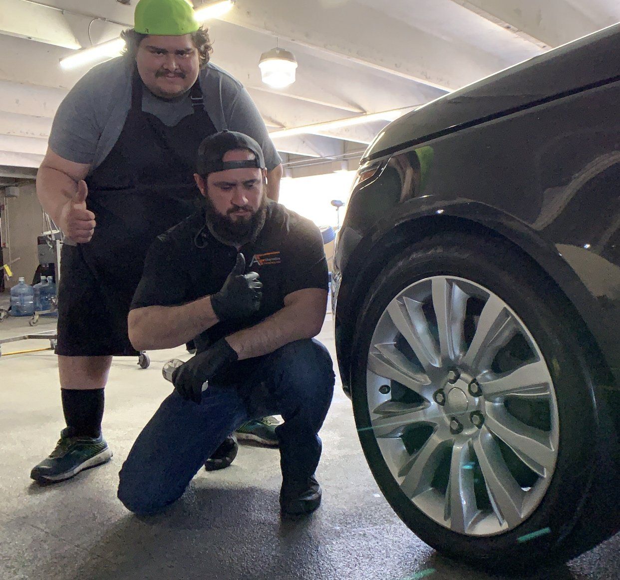 Two men pose with a black car. One kneels by the tire, holding a tool. The other gives a thumbs up and wears a green hat.