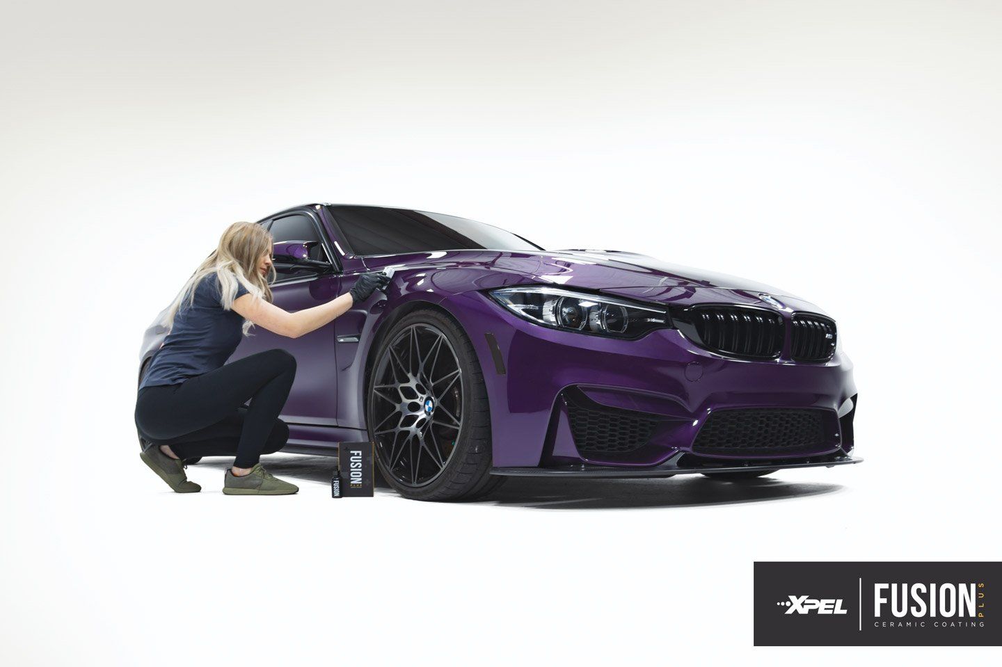 Woman applying coating to a purple BMW car. White background, black wheels, XPEL Fusion logo.