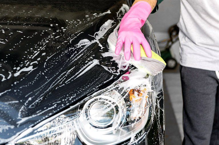 Person washing a black car with soapy water and a sponge, wearing pink gloves.