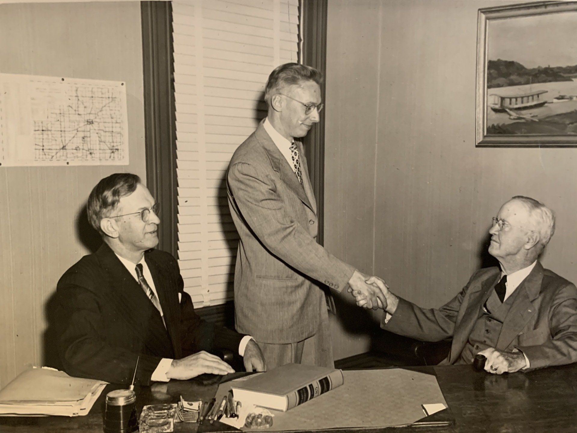Three men in suits shaking hands in an office; a map and painting hang on the wall.