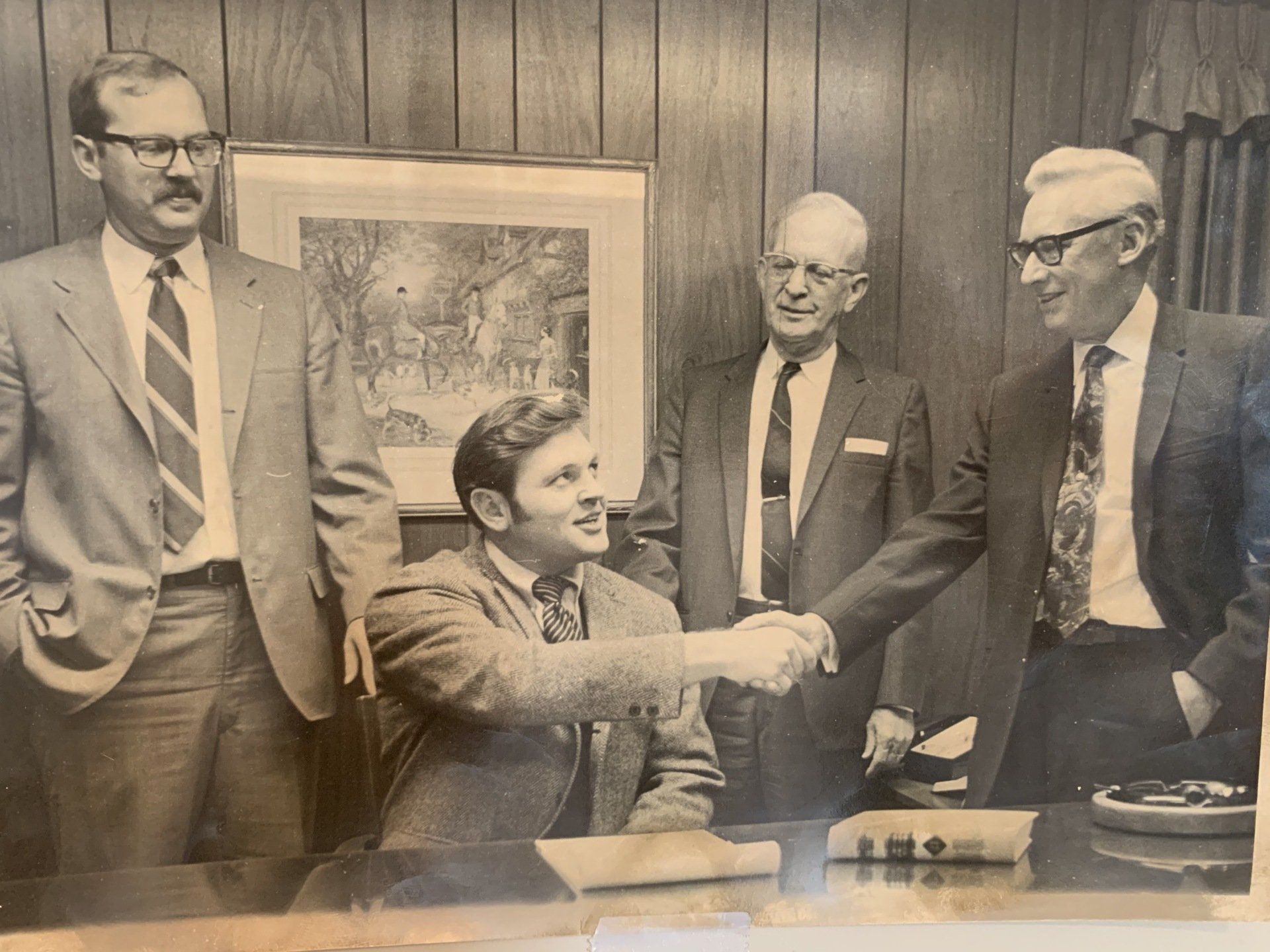 Four men in suits, one seated shaking hands, standing near a wooden wall.