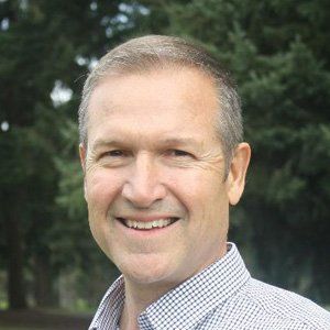 Man with short, graying hair and a button-down shirt smiles in front of trees.