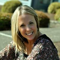 Blond woman smiles, outdoors. Floral print top, setting with green bushes.