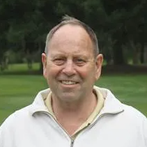 Man smiling outdoors, wearing a white zip-up sweater. Green grass and trees in background.