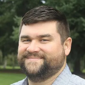 Man with a beard, smiling, outdoors in front of a tree.