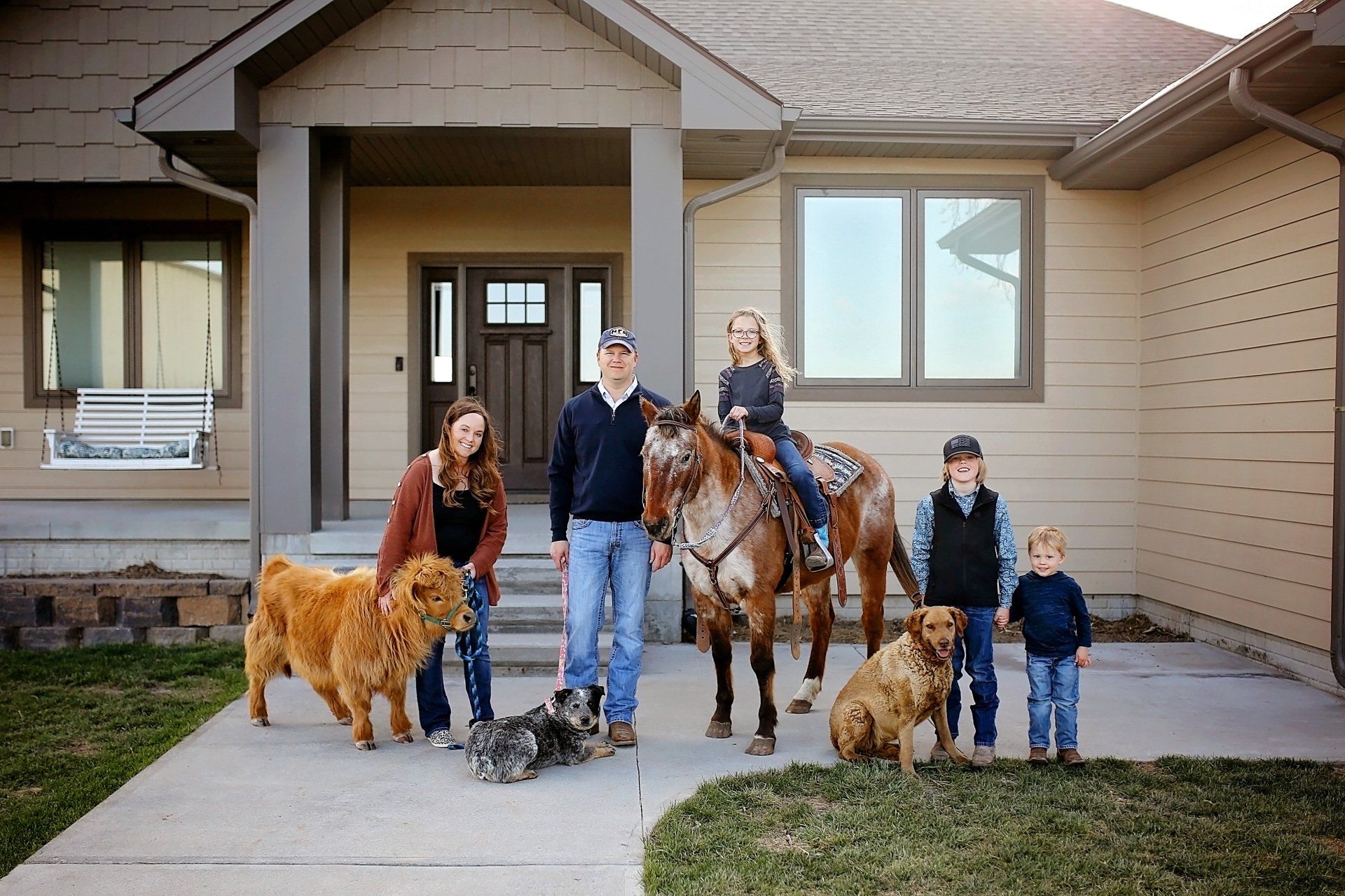 Family and pets pose in front of a house. Woman and girl on horse. Others with dogs.