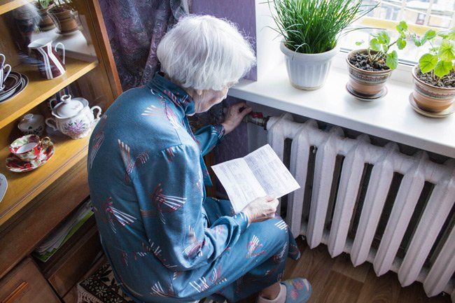 An elderly woman is sitting on a radiator reading a book.