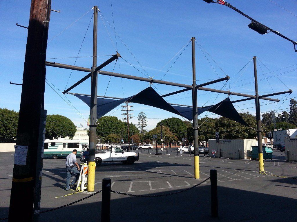 Parking lot with shade sails, white car, and a few people near a street intersection
