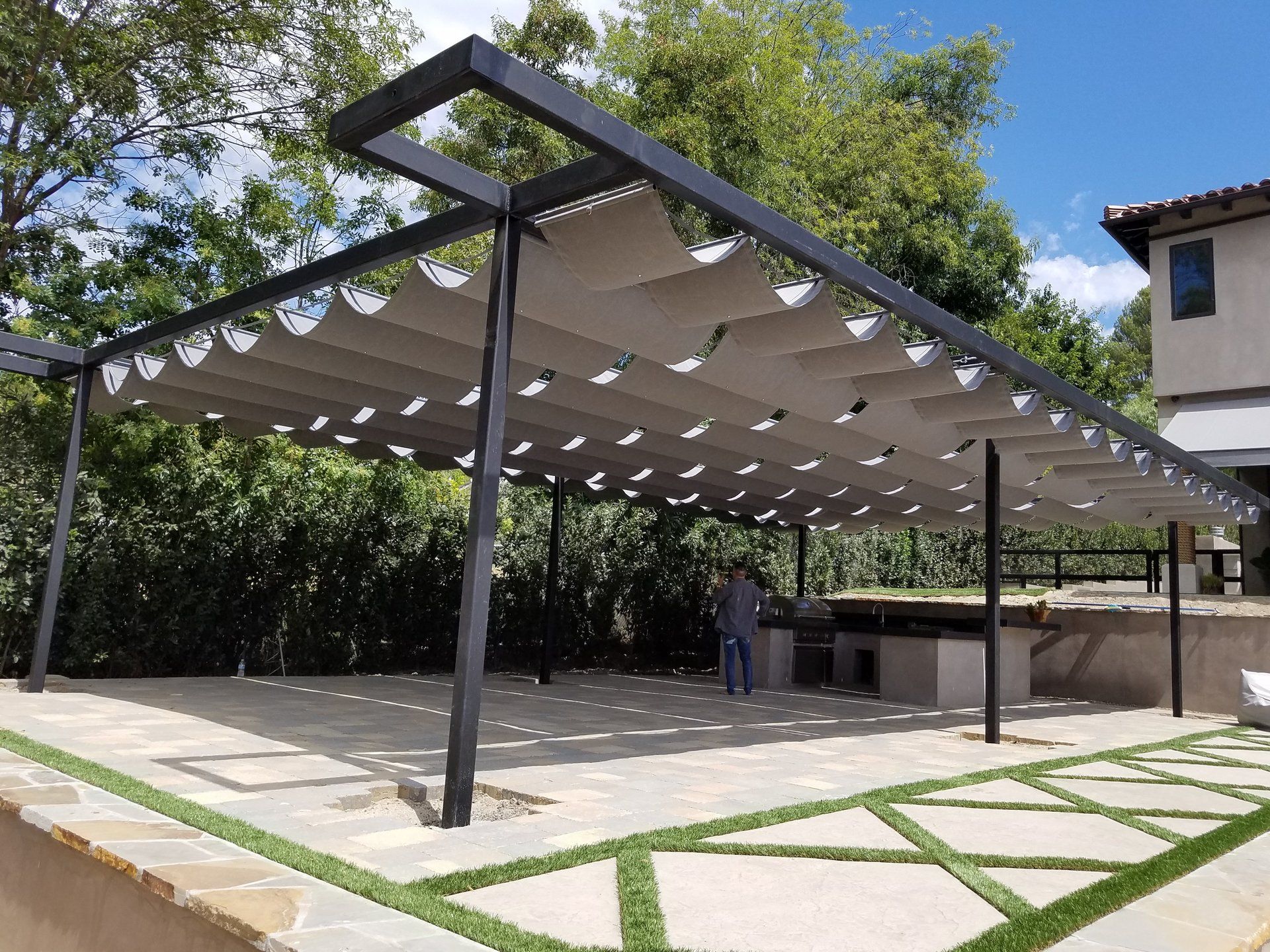 Modern outdoor pergola with black frame and white shade sails over a paved patio in a garden area
