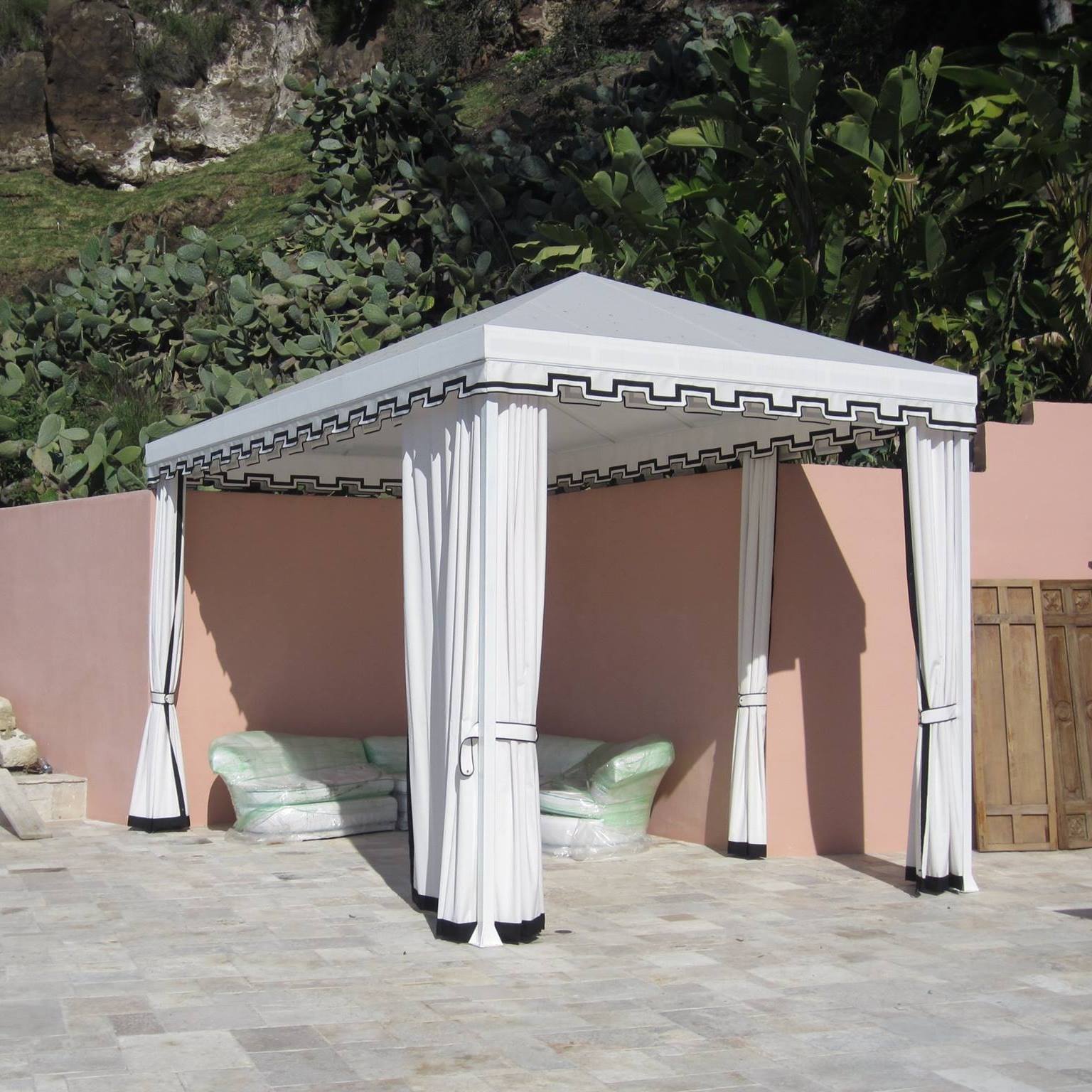 White gazebo with curtains and green cushions on a stone patio beside a pink wall and garden greenery