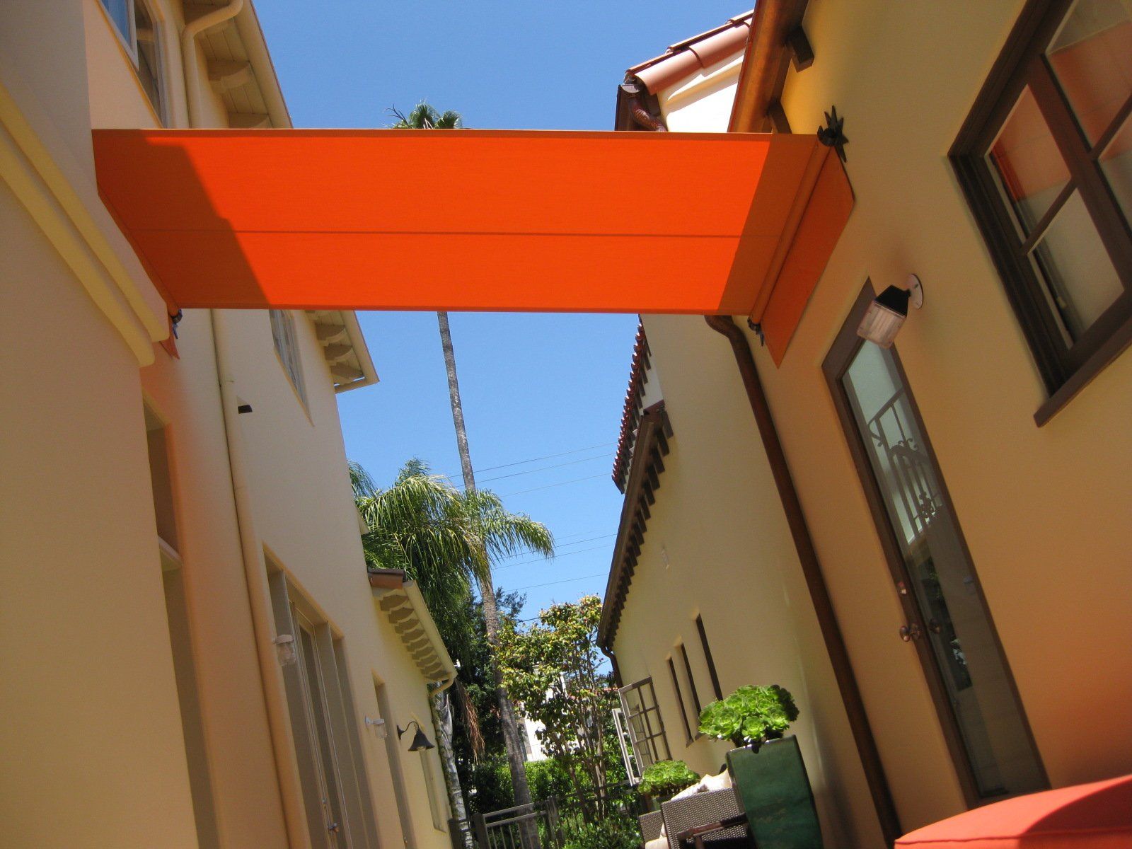 Narrow alley between cream buildings, linked by a bright orange overhead bridge under a blue sky.