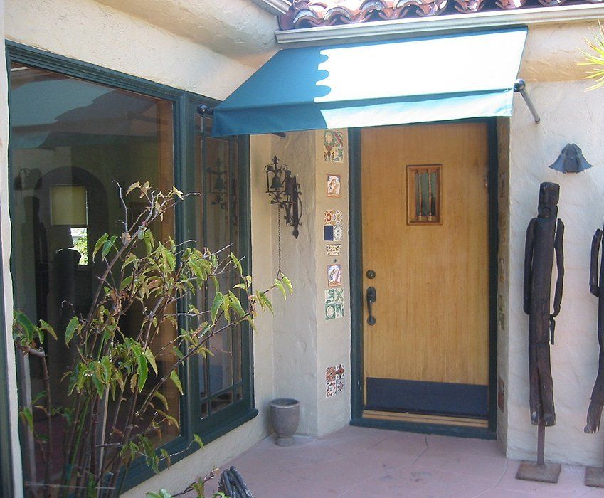 Front entryway with a blue awning, wooden door, potted plant, and narrow courtyard.