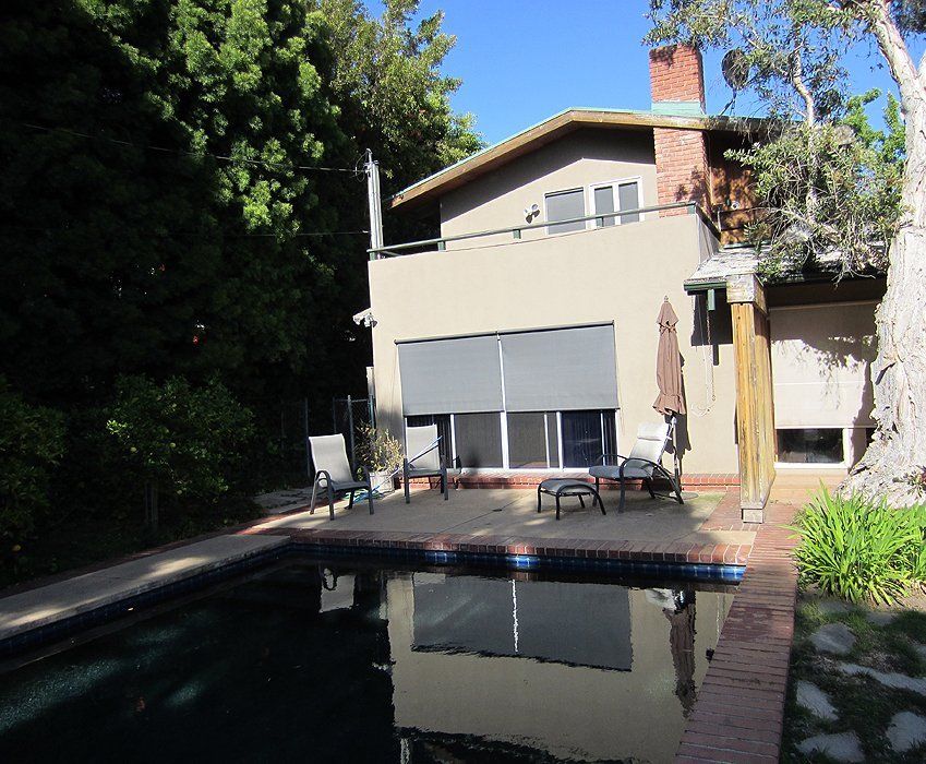 Backyard pool beside a house with patio chairs, brick path, and trees in shade