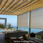 Beachfront patio with shaded lounge chairs, a table, and ocean view behind bamboo blinds.