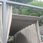 Covered outdoor walkway beside a white wall and curtains, with trees visible above.