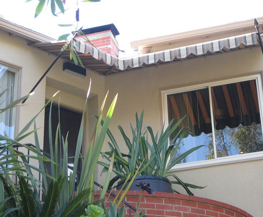 Courtyard view of beige house exterior with window, striped awning, and tall green plants in foreground