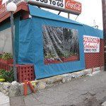 Blue tarp-covered roadside stall with a Coca-Cola sign and small potted plant on a concrete corner