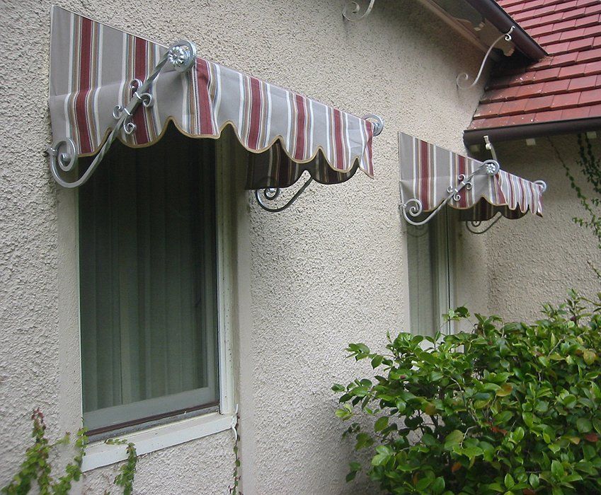 Two striped window awnings on a stucco house above a window and shrubbery.