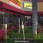 Red storefront with yellow awning, palm tree, and patio enclosure along a sidewalk.