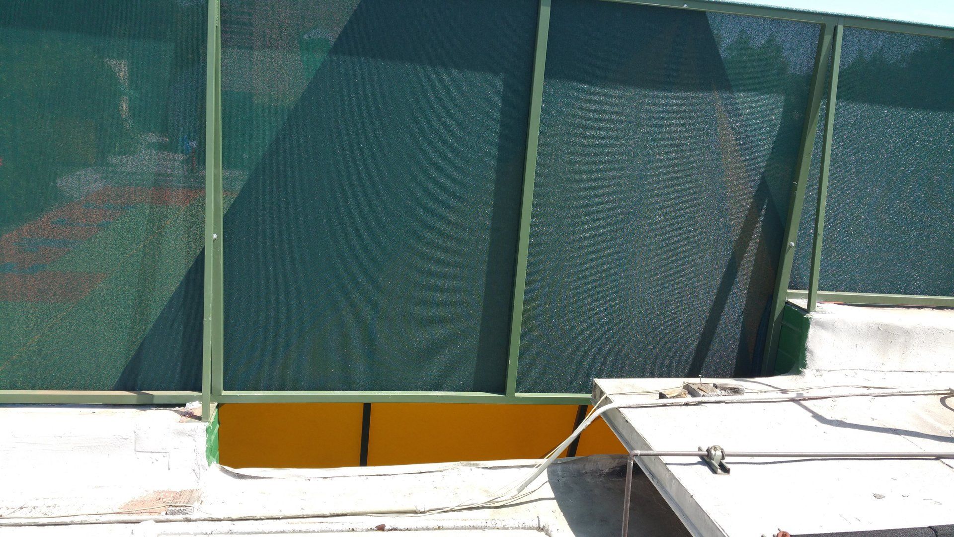 Green mesh fencing above a white rooftop with a yellow wall panel and metal railing.