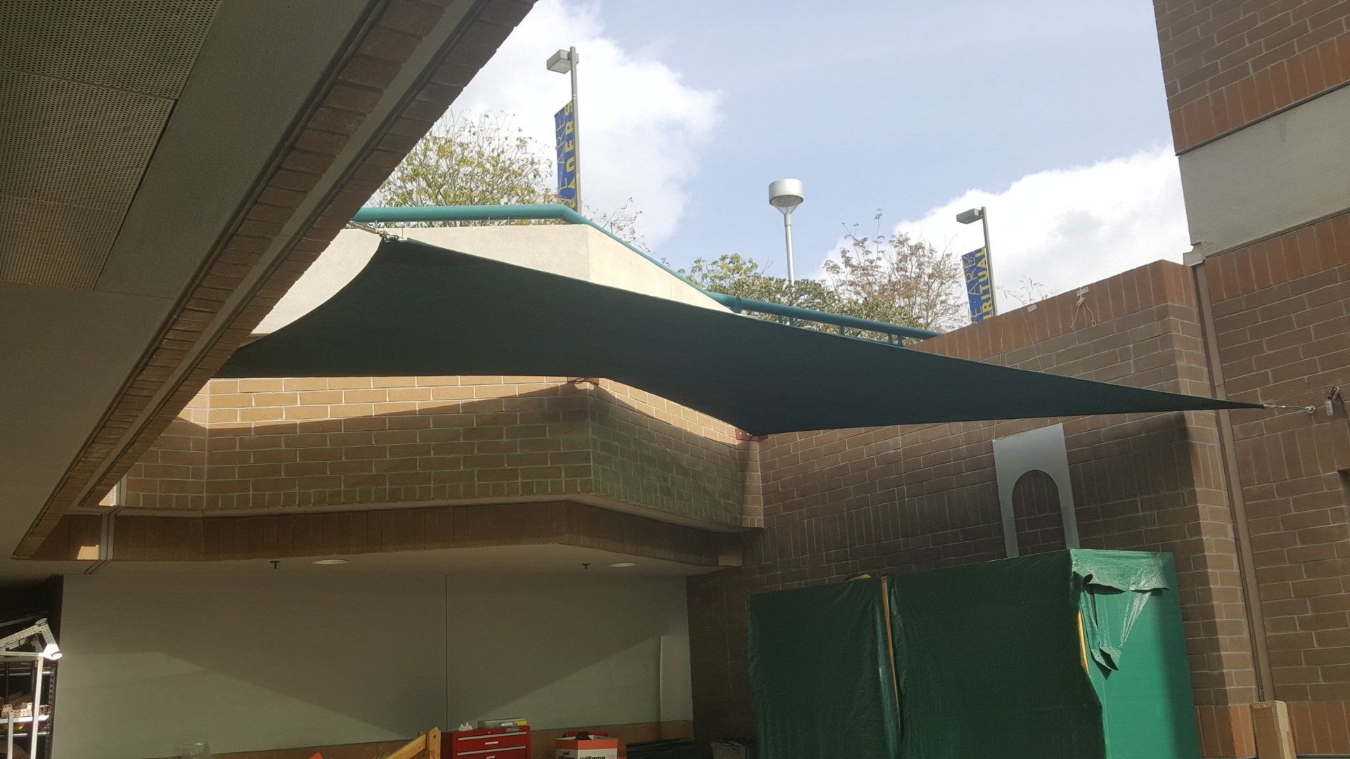 Shaded courtyard with green canopies beside brick buildings and a green gate under a partly cloudy sky