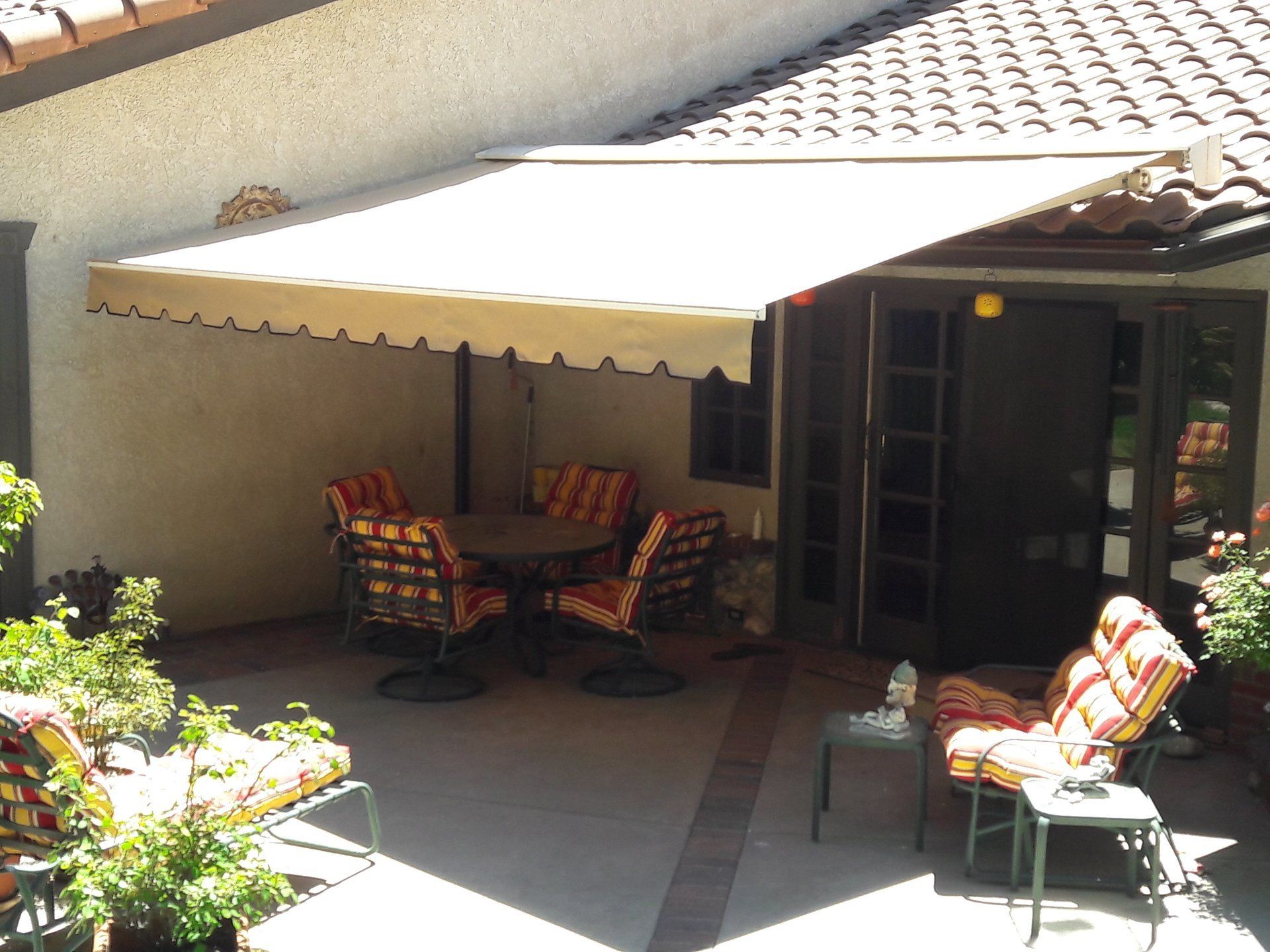 Patio with white awning, small table and patterned chairs, surrounded by potted plants and a glass door