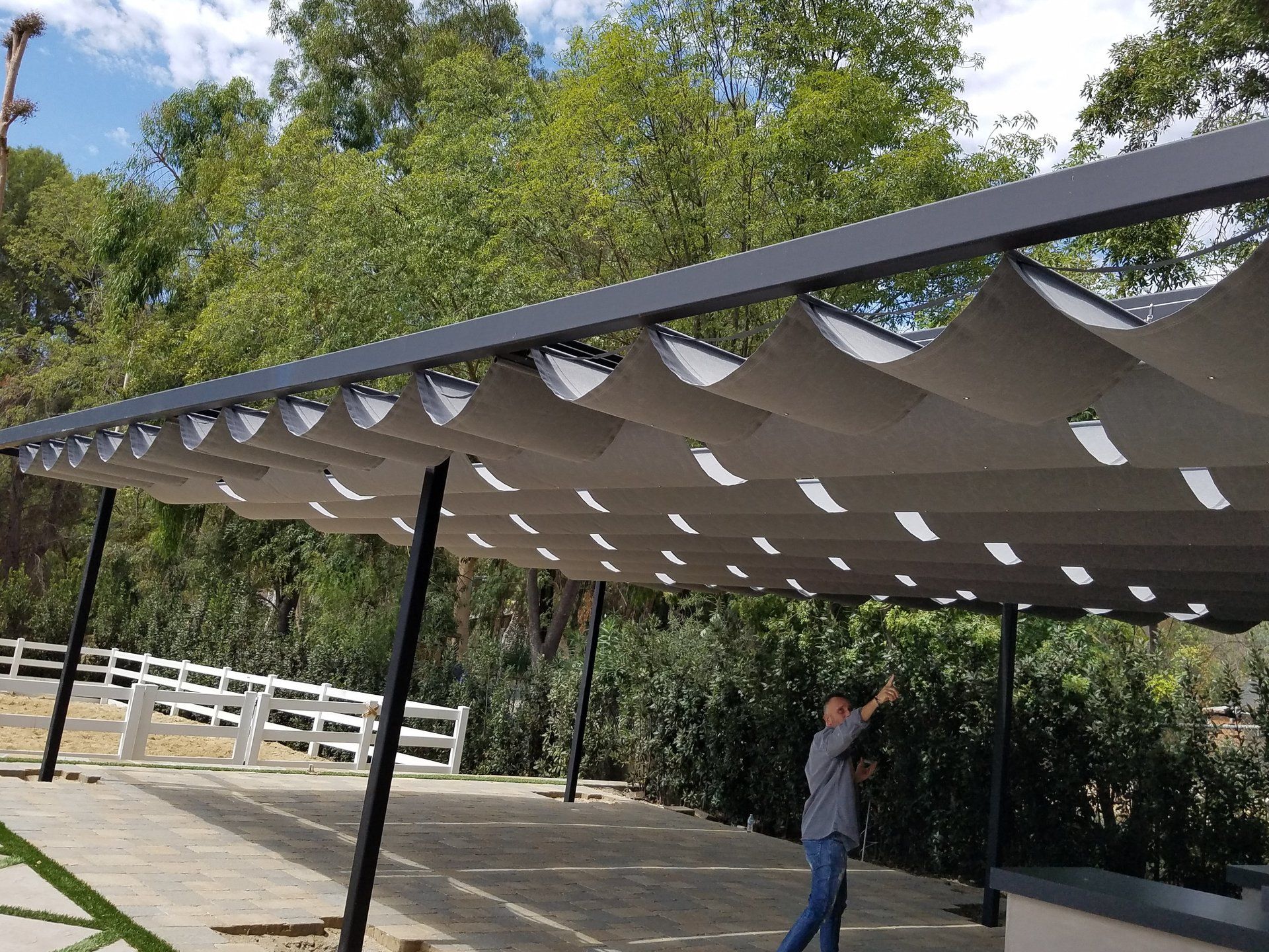 Person standing under a large shaded canopy in a park with trees and white railings nearby
