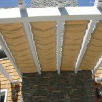 Close-up of beige scalloped awnings hanging from a white pergola over a stone patio.