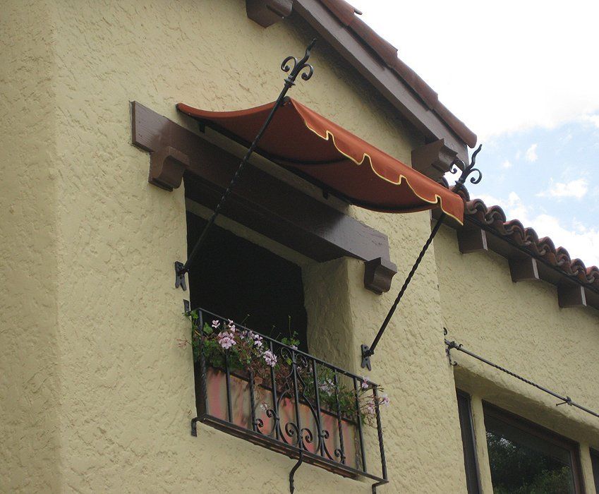 Small balcony with flower box and wrought-iron railing under a red fabric awning on a stucco house