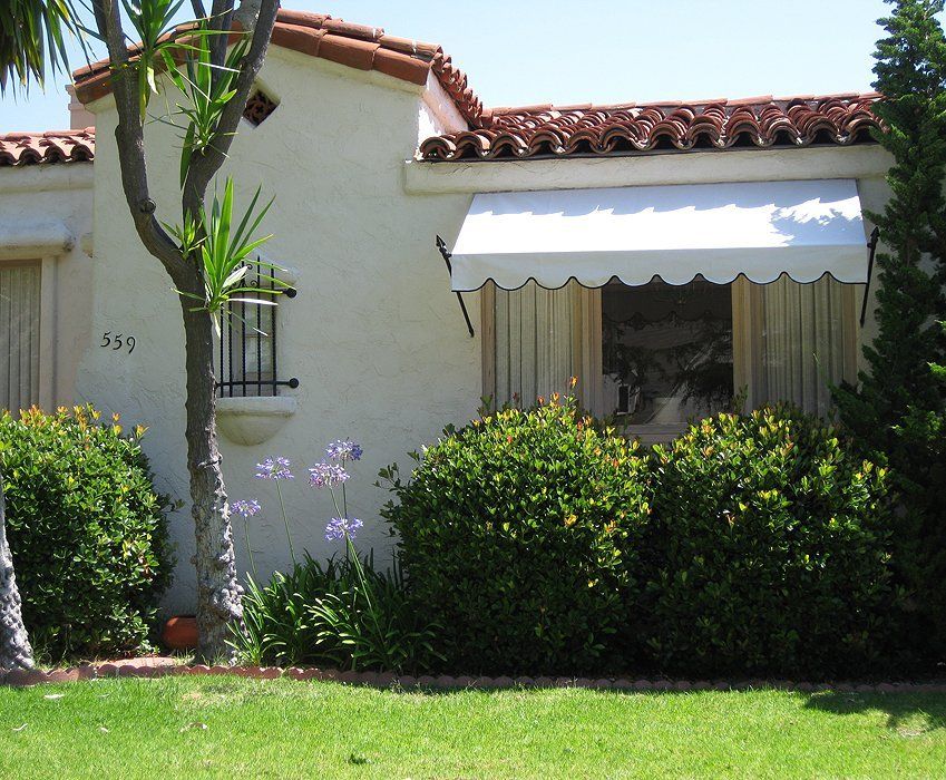 Small stucco house with red tile roof, white awning, and lush green shrubs in front yard