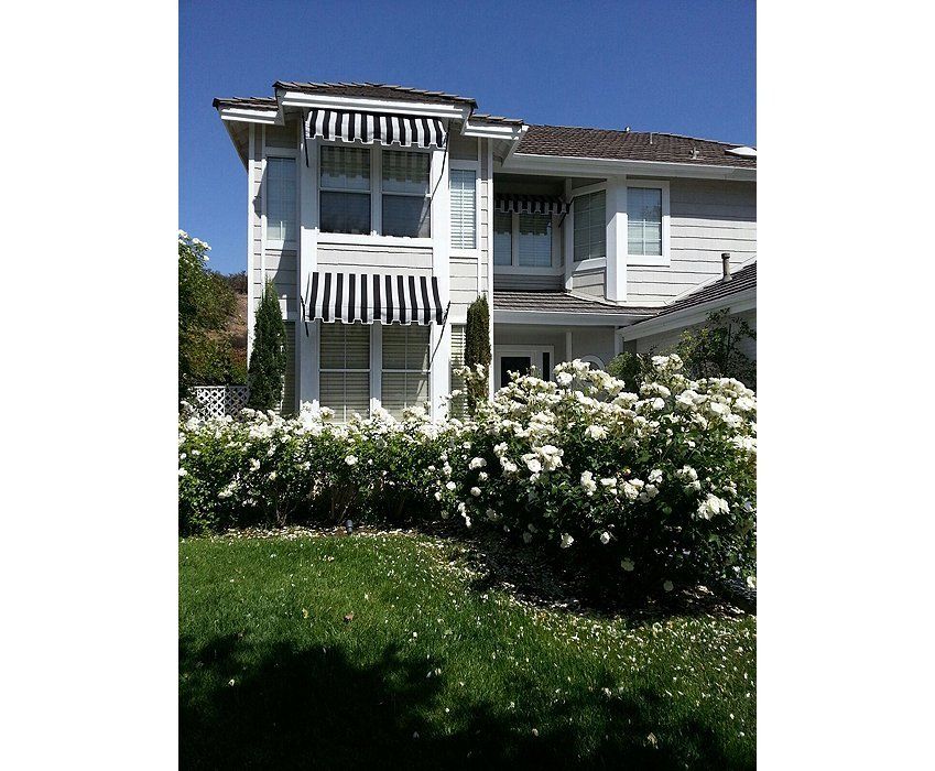 Two-story white house with striped awnings, flowering bushes, and green lawn under a blue sky