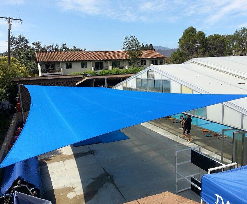 Blue shade sail over outdoor courtyard with buildings and trees in the background