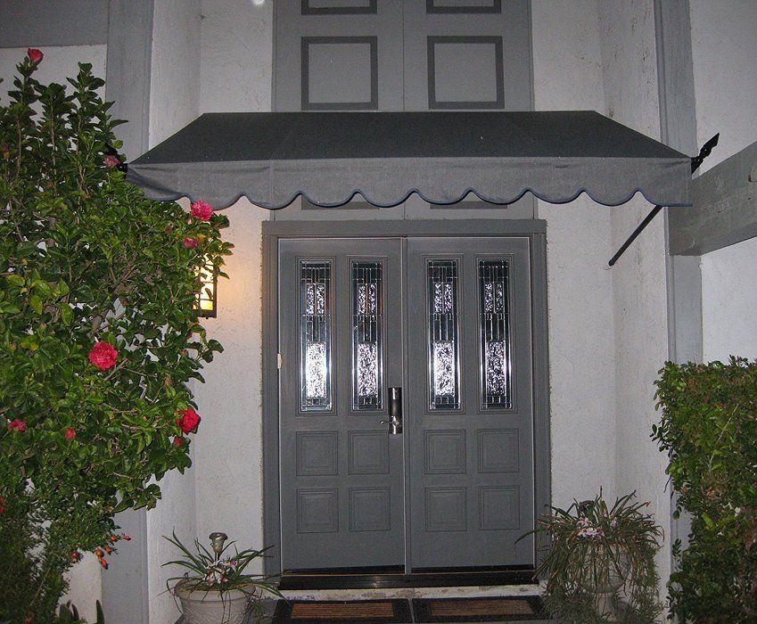 Gray double front doors with a scalloped awning, framed by flowering shrubs at night