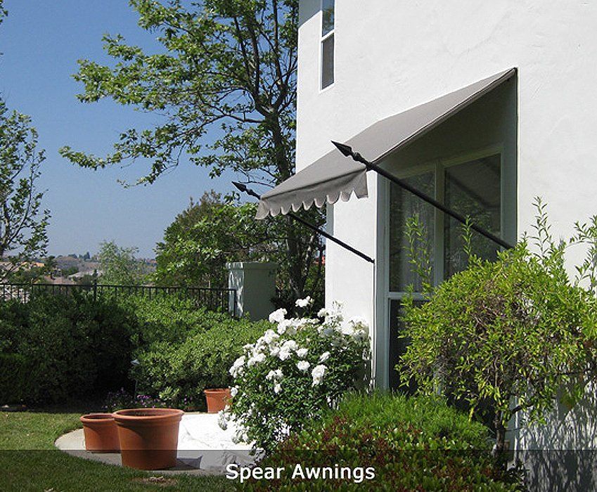 White house with black spear awning shading a window, surrounded by green shrubs and patio pots.