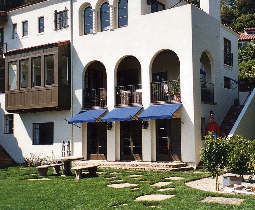 White stucco building with blue awnings, arched balconies, and a grassy front yard with stepping stones