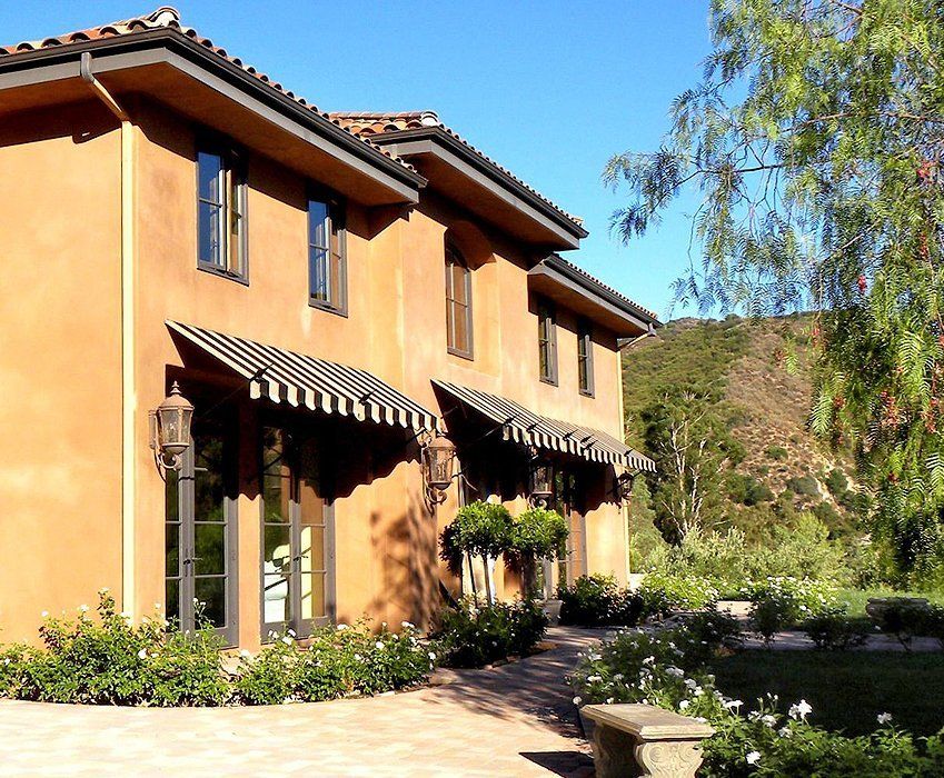 Sunlit stucco house with striped awnings, garden path, and trees against a hillside