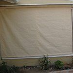 Closed beige outdoor shade covering a patio wall with plants at the base