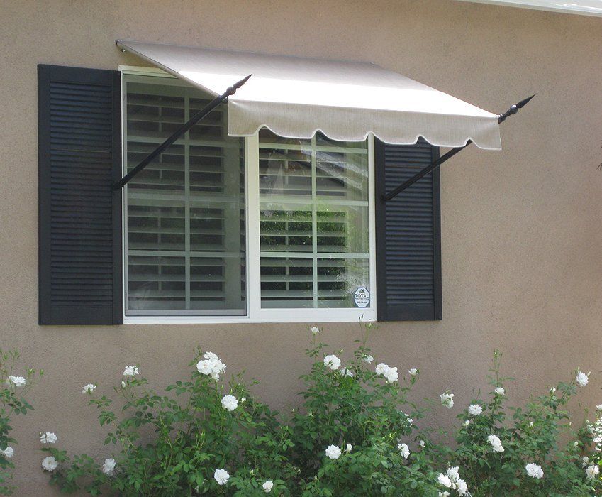 Window with dark shutters and a white awning above flowering bushes on a beige house exterior