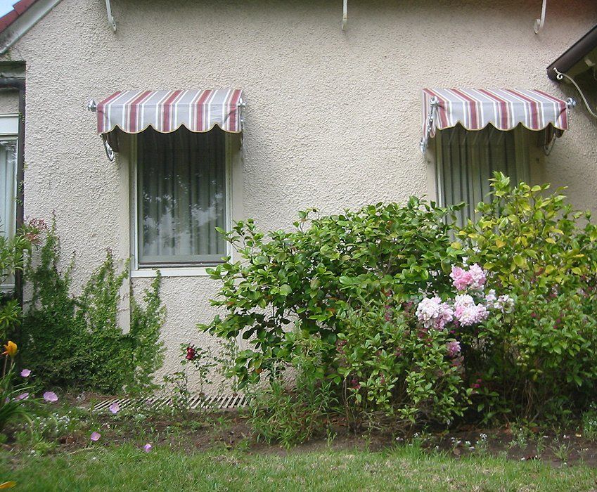 Small house facade with striped awnings, windows, and a garden with pink flowers and green shrubs.